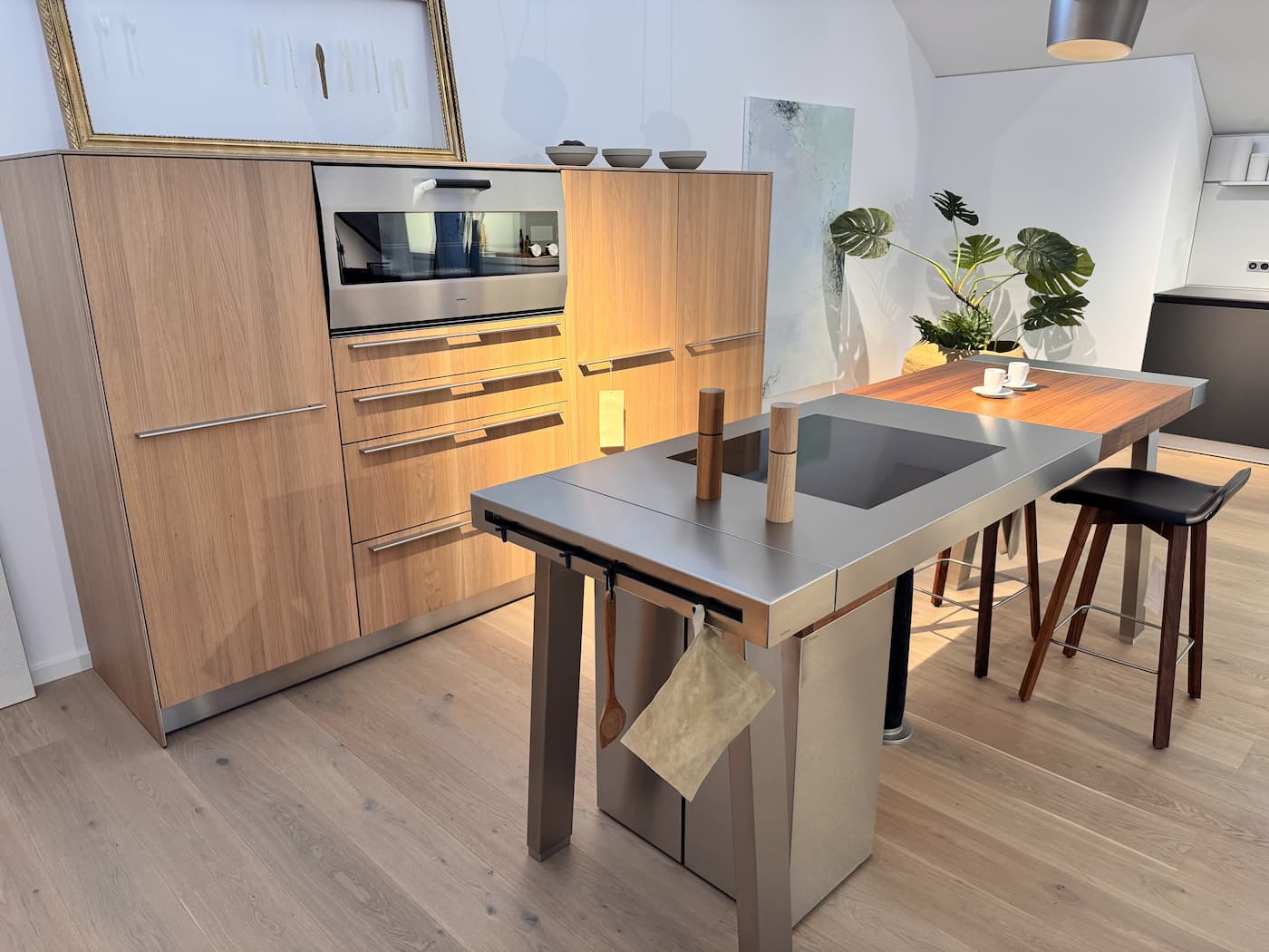 bulthaup Kochinsel aus Edelstahl mit Holztisch, dahinter Hochschrank in hellem Holz mit Backofen